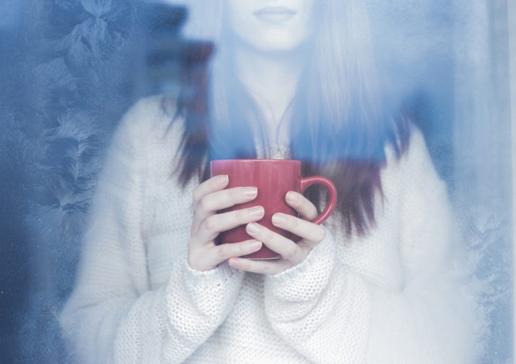 Woman in white sweater looking our frosted window, holding red mug.