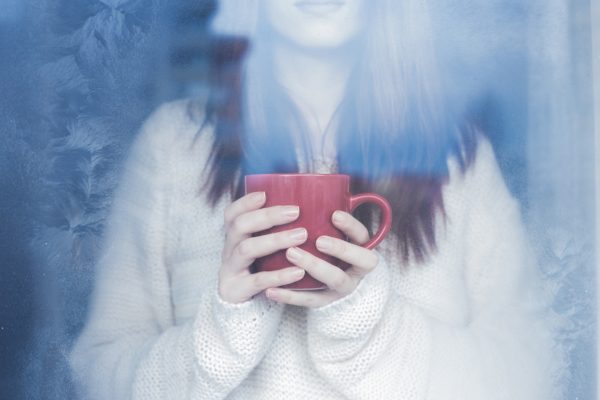 Woman in white sweater looking our frosted window, holding red mug.