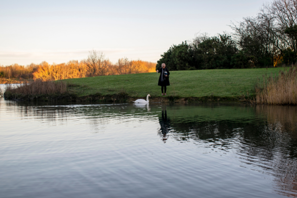 Wide shot of pond with woman dressed in winter clothes, standing on the edge of field, holding urn. Swan is in pond, close to woman.