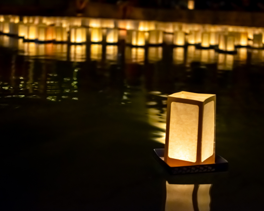 Lantern floating on dark water with dozens of lanterns also floating on water in background.