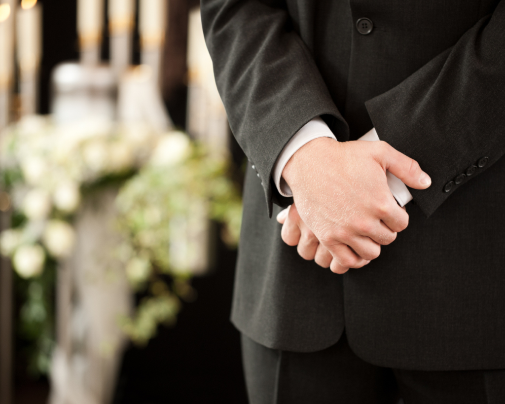 Close up of funeral directors hands crossed, wearing dark suit. Floral arrangements can candles can be seen in the background.