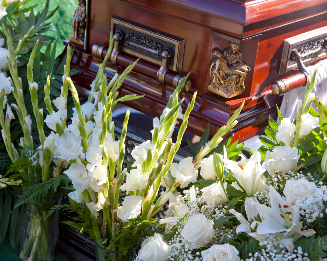 Casket surrounded by white roses and lilies outdoors, with sunlight beaming down on it.