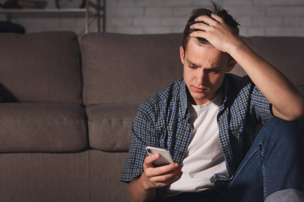 A person sitting on a couch, looking at their phone with a sad expression while holding their head.