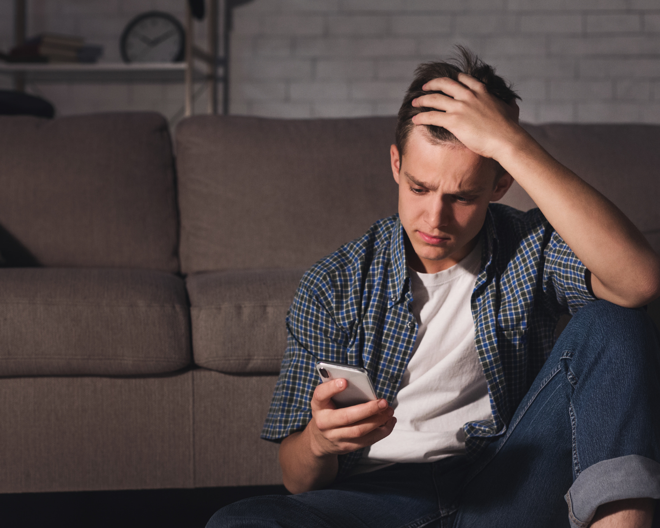 A person sitting on a couch, looking at their phone with a sad expression while holding their head.