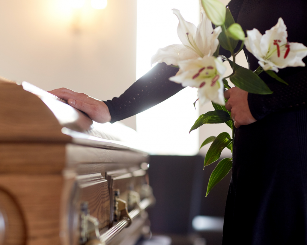 A person holding white lilies rests a hand on a casket during a funeral service, symbolizing remembrance and the funeral process.