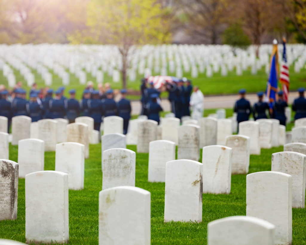 Military funeral ceremony at a cemetery with uniformed service members standing around a gravesite holding the American flag and the U.S. Navy flag.