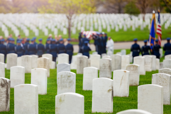 Military funeral ceremony at a cemetery with uniformed service members standing around a gravesite holding the American flag and the U.S. Navy flag.