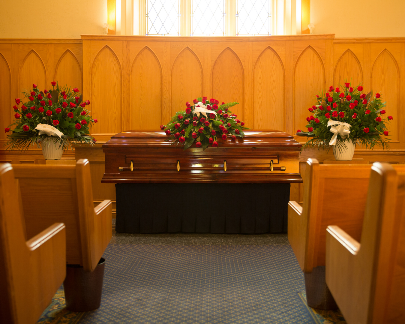A polished dark wood casket adorned with red roses sits at the front of a chapel between church pews, flanked by two matching red rose floral arrangements against a wooden paneled wall.