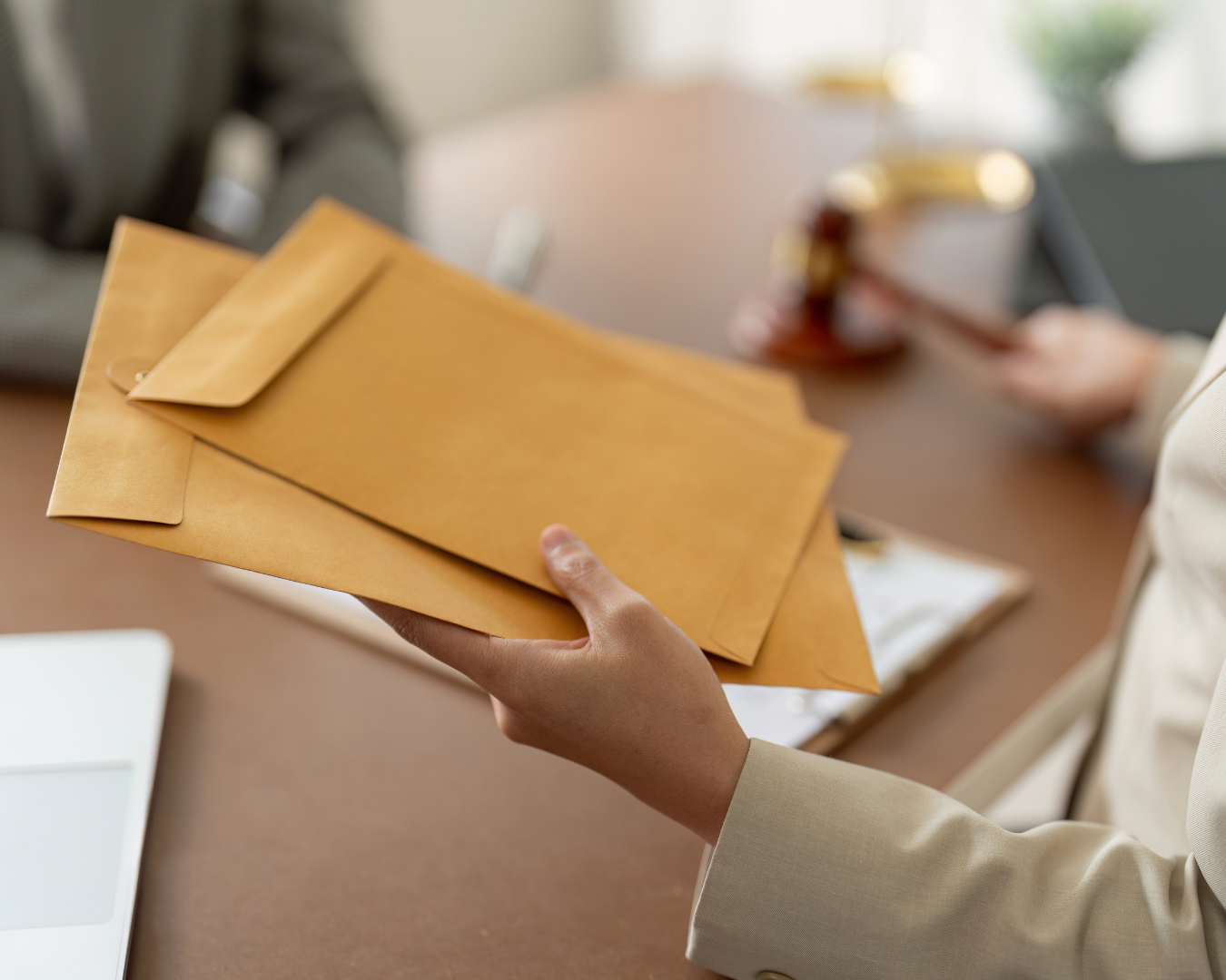A close-up shot focusing on a person in a beige blazer holding two manila envelopes over a wooden desk in a legal setting. In the blurred background, another individual in a suit sits across the desk, and a legal gavel is visible.